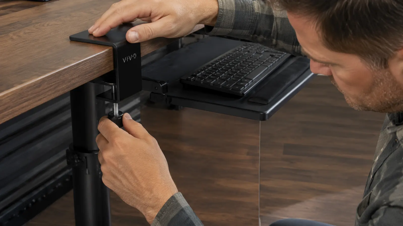 Close-up of a hand tightening the C-clamp mount of a black VIVO keyboard tray to the underside of a desk