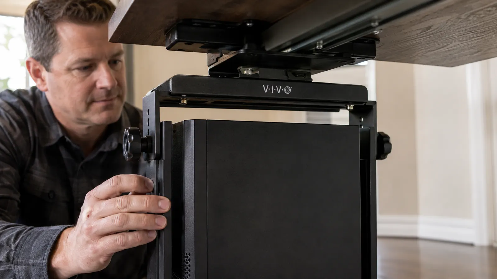 Close-up of a hand tightening the adjustable side clamps on a black VIVO under-desk CPU mount holding a computer tower