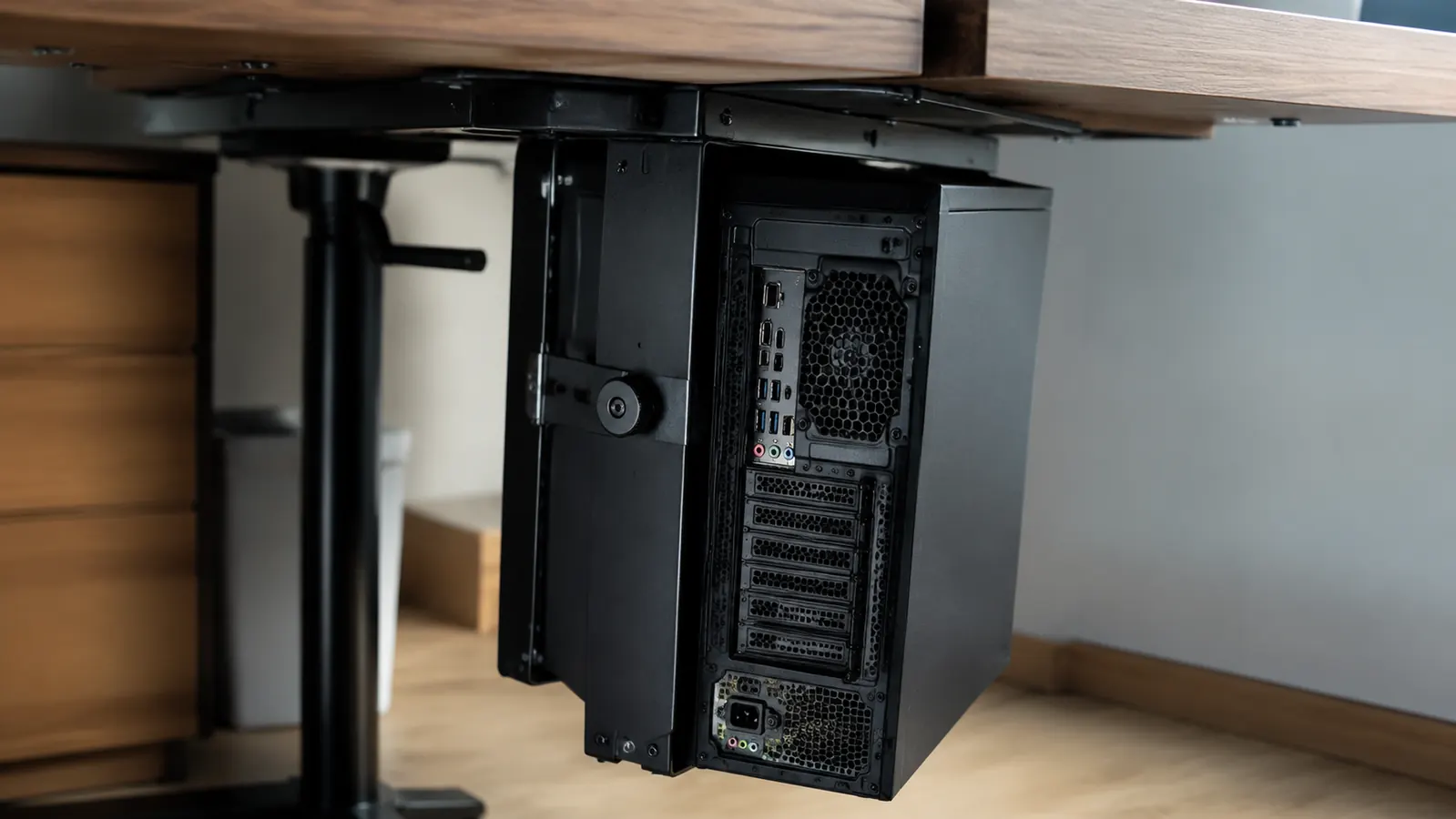 Close-up of a steel CPU mount bracket clamped to the underside of a desk with a tower case swiveled outward