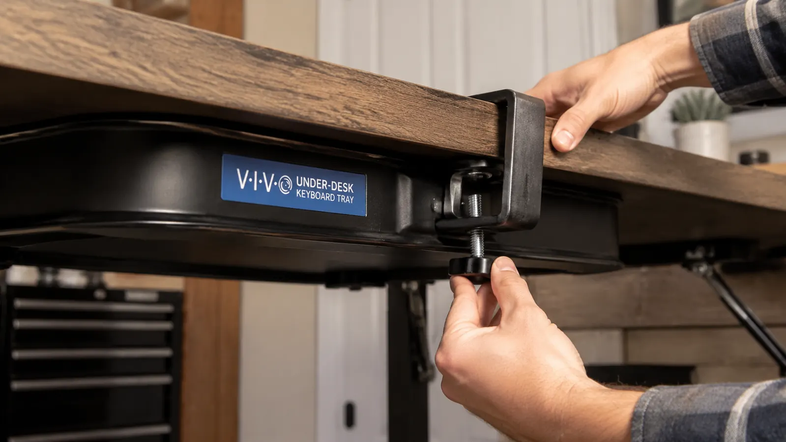 Close-up of a hand tightening the C-clamp mount of a black VIVO keyboard tray to the underside of a desk